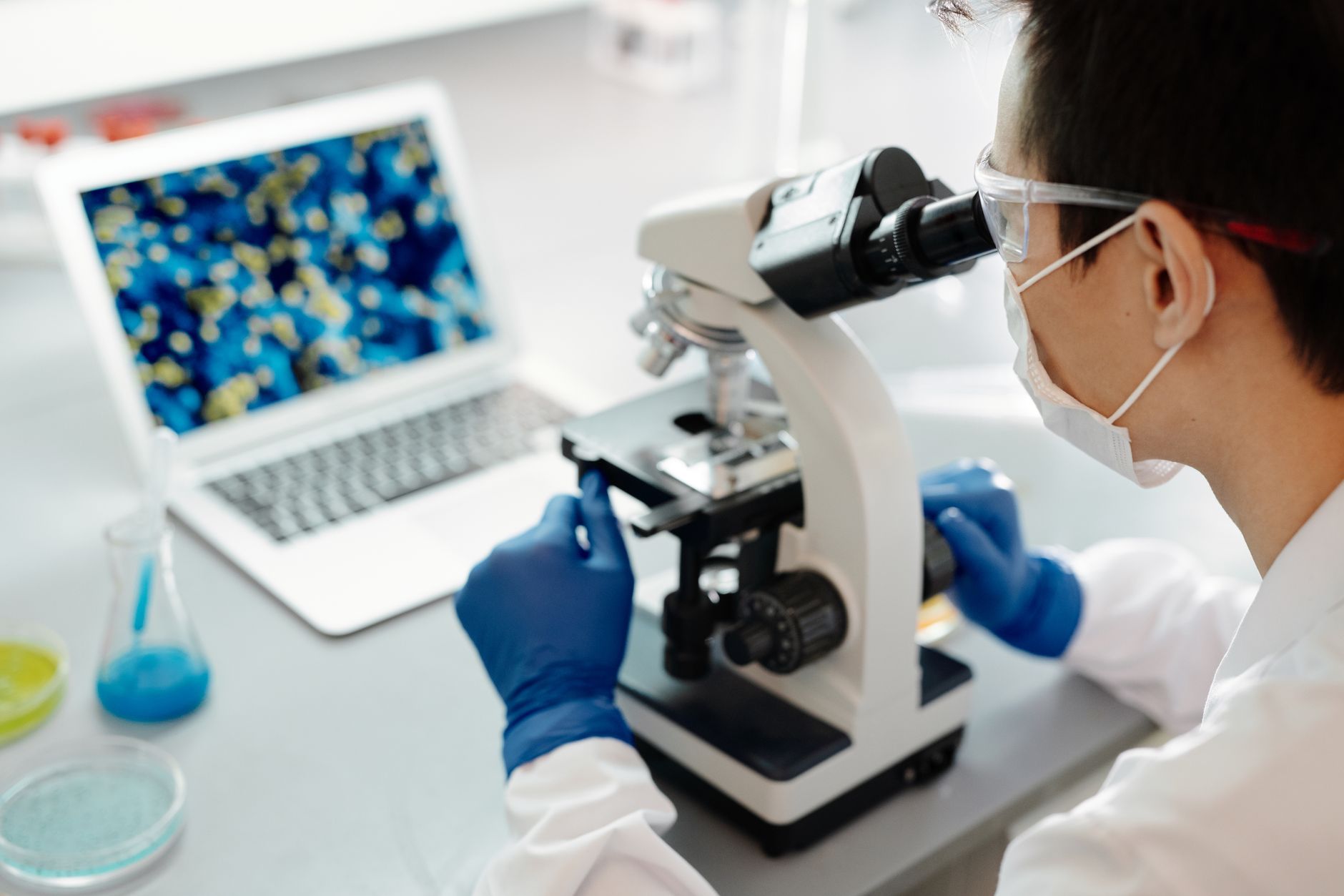 Forscher mit Schutzbrille, Maske und blauen Handschuhen am Mikroskop, daneben ein Laptop mit einer mikroskopischen Zellaufnahme