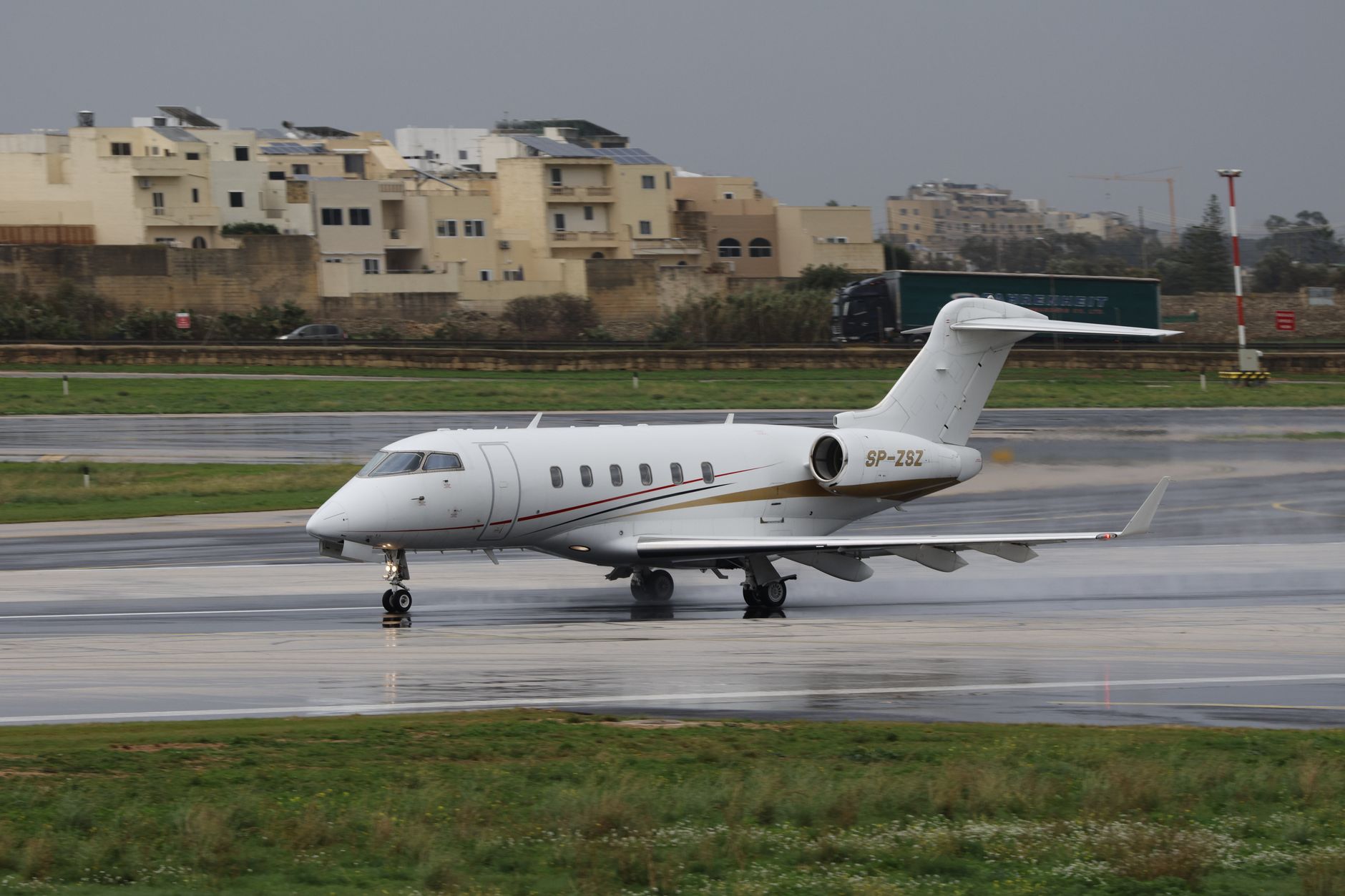 Weißes Privatjet-Flugzeug auf nasser Piste, im Hintergrund sandfarbene Gebäude, Grünstreifen und grauer Himmel.